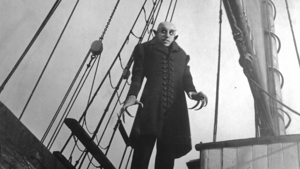 A scene from Nosferatu, the main character Max Schreck stands in front of the rigging of a sailing ship and looks down ominously.